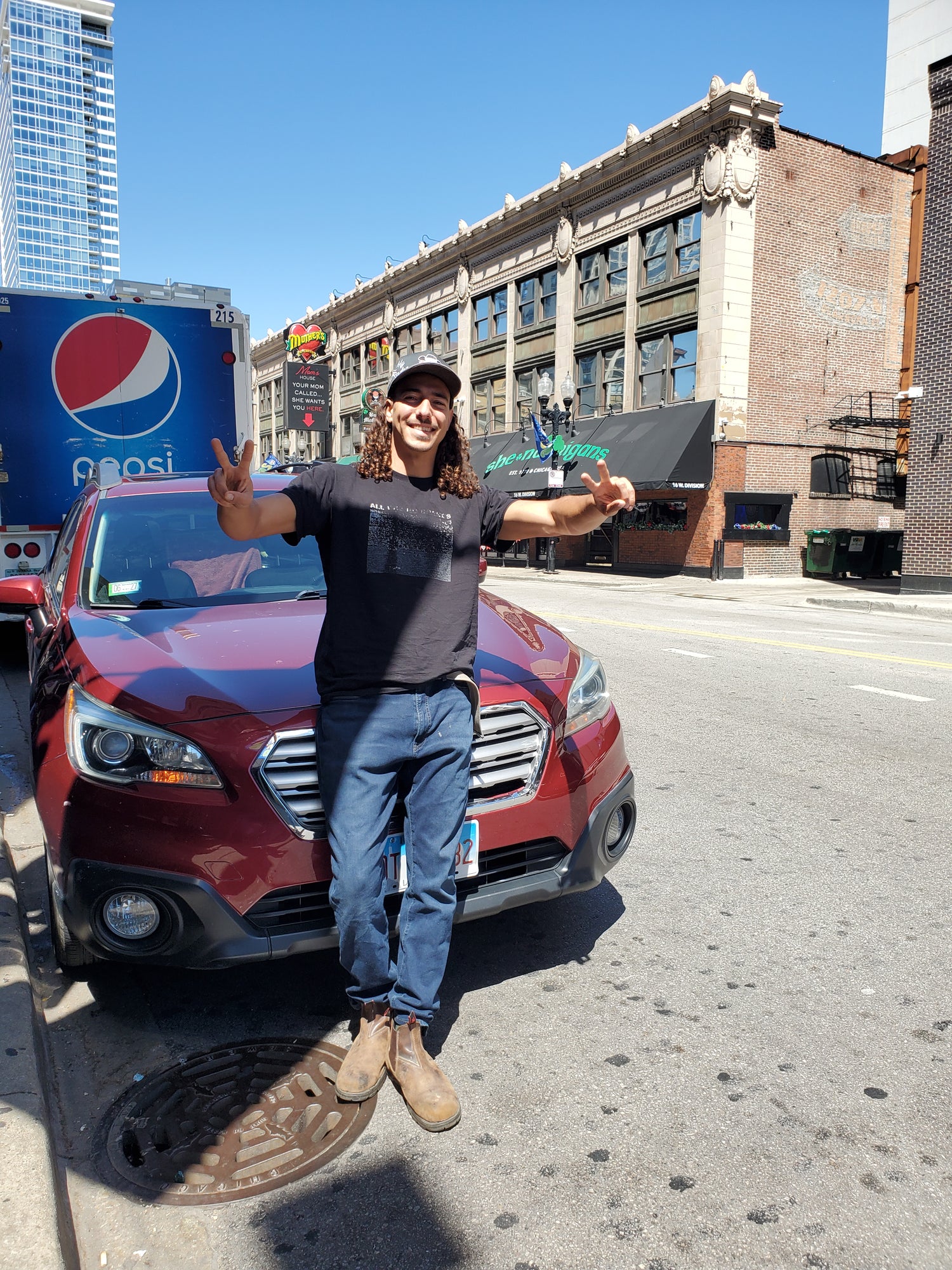 A man wearing a black t-shirt, blue jeans, and brown boots stands in front of a red SUV on a city street, gesturing with peace signs. A blue Pepsi truck is visible on the left, and brick buildings line the street under a clear blue sky.